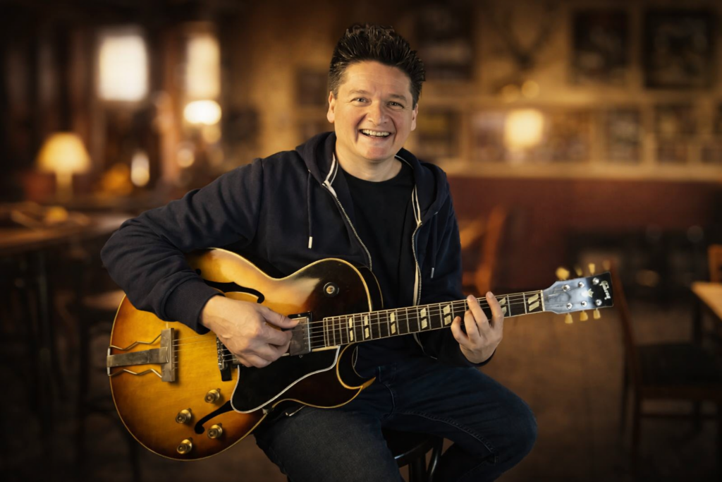 Porträtfoto eines lächelnden Mannes, der auf einem Stuhl sitzt und eine elektrische Jazzgitarre hält. Er trägt dunkle Kleidung und blickt direkt in die Kamera. Die Gitarre hat eine warme Holzfarbe und liegt spielbereit auf seinem Bein. Der Raum wirkt wie ein Café oder Proberaum mit Tischen und Stühlen im Hintergrund, der unscharf dargestellt ist. Das Licht ist warm und erzeugt eine freundliche, entspannte Atmosphäre.